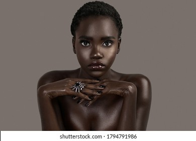 A portrait of a serene young black female with short black hair, moist lips, perfectly black manicured nails with silver jewelry looking straight at the camera.
