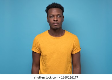 Portrait Of Serious African American Man In Yellow Shirt With No Emotion On Face Against Blue Background.