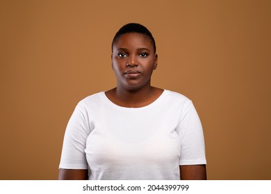 Portrait Of Serious African American Young Woman Wearing White T-Shirt Posing Looking At Camera Standing Isolated Over Brown Studio Background. Female Headshot Of Black Lady