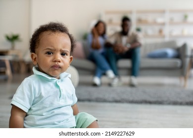 Portrait Of Serious Angry African American Baby Boy Looking At Camera, Funny Adorable Toddler Sitting On The Floor, Parents Resting On Sofa In The Blurred Background, Selective Focus On Little Kid