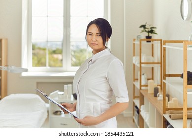 Portrait Of Serious Asian Cosmetologist Or Aesthetic Nurse In Her Workplace. Professional Beautician With Clipboard Standing In Beauty Salon. Female Massagist In Massage Room Looking At Camera