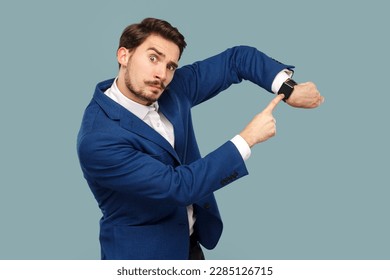 Portrait of serious assertive man with mustache standing pointing at his wristwatch, saying you have no time, wearing white shirt and jacket. Indoor studio shot isolated on light blue background.