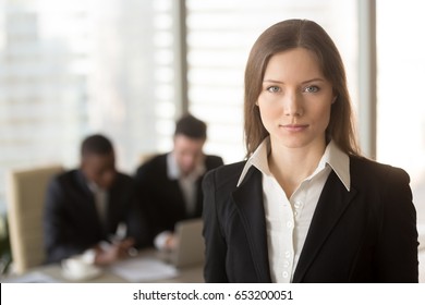 Portrait of serious attractive businesslady in office interior with male colleagues at the background, looking at camera, ambitious motivated career woman, successful female boss running own business