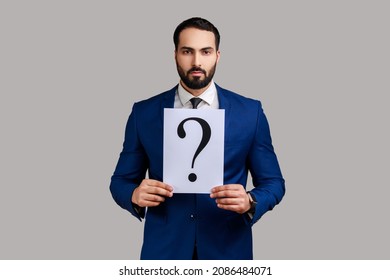 Portrait of serious bearded businessman looking at camera, holding paper with question mark, thinks about tasks, wearing official style suit. Indoor studio shot isolated on gray background. - Powered by Shutterstock