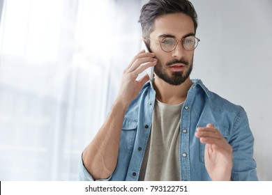 Portrait Of Serious Bearded Male Wears Glasses And Fashionable Denim Shirt, Has Phone Conversation, Gestures With Hand As Tries To Explain Something, Solves Problems Via Smart Phone On Distance