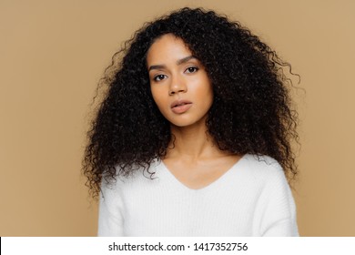 Portrait Of Serious Beautiful Dark Skinned Female With Frizzy Black Hair, Has Minimal Makeup, Looks Calmly At Camera, Wears White Jumper, Stands Against Brown Background, Being Deep In Thoughts.