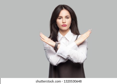 Portrait Of Serious Beautiful Young Woman In White Shirt And Brown Apron With Makeup And Brunette Hair Standing And Showing X Sign And Looking At Camera. Studio Shot, Isolated On Grey Background.