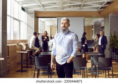 Portrait Of Serious Business Teacher And Professional Team Coach. Handsome Man With White Hair And Grey Beard Standing In Office After Corporate Training Class, With Group Of Employees In Background