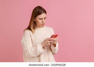 Portrait Of Serious Concentrated Attractive Blond Woman Using Mobile Phone, Looking At Device Screen, Typing Message, Wearing White Sweater. Indoor Studio Shot Isolated On Pink Background.