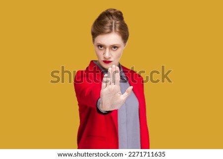Portrait of serious concentrated woman with red lips showing stop gesture with palm, looking at camera, protecting herself, wearing red jacket. Indoor studio shot isolated on yellow background.
