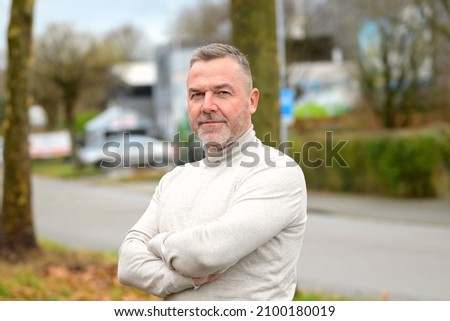 Portrait of an serious confident middle-aged man with enigmatic expression standing in a quiet city street looking intently at the camera with folded arms