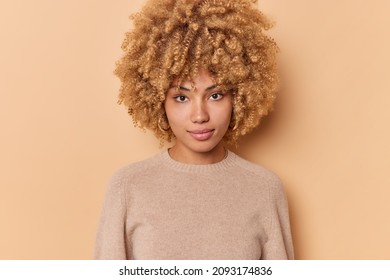 Portrait of serious confident sassy good looking woman with bushy curly hair looks directly at camera stands indoor against beige background wears casual jumper. Human face expressions concept