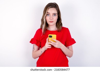 Portrait Of Serious Confident Young Caucasian Woman Wearing Red T-shirt Over White Background Holding Phone In Two Hands