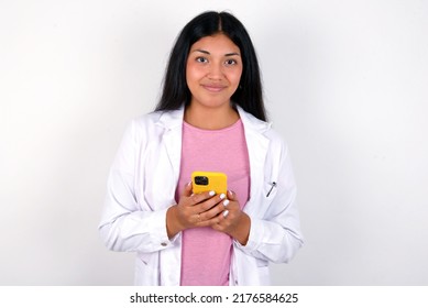 Portrait Of Serious Confident Young Hispanic Doctor Girl Wearing Coat Over White Background Holding Phone In Two Hands