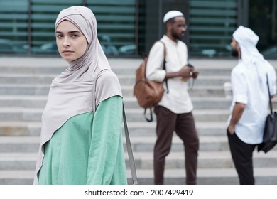 Portrait Of Serious Confident Young Muslim Woman In National Clothes Wearing Shoulder Bag Against Colleagues Outdoors