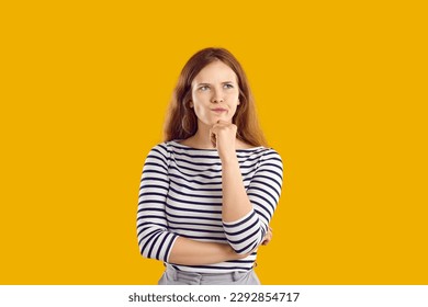 Portrait of serious confused undecided young woman full of doubt holding hand on chin and thinking, trying to make hard choice decision. Girl with wavy hair in striped sweatshirt on yellow background.