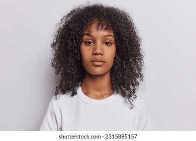 Portrait of serious curly haired woman looks directly at camera has attenitve gaze calm expression dressed in casual t shirt isolated over white background. Pretty millennial girl poses in studio - Shutterstock ID 2315885157