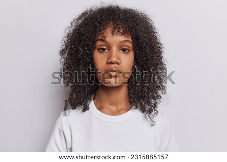 Portrait of serious curly haired woman looks directly at camera has attenitve gaze calm expression dressed in casual t shirt isolated over white background. Pretty millennial girl poses in studio