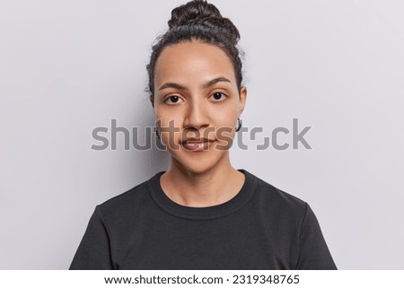 Portrait of serious dark haired young Latin woman has healthy well cared skin poses without makeup has attentive gaze at camera dressed in casual black t shirt isolated over white background