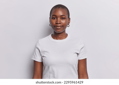 Portrait of serious dark skinned woman with neutral facial expression looks directly at camera dressed in casual t shirt and golden earrings isolated over white background. Ethnicity concept - Shutterstock ID 2295961429