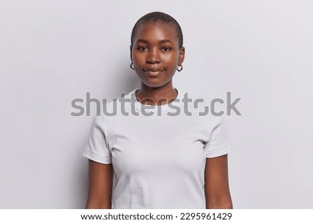 Portrait of serious dark skinned woman with neutral facial expression looks directly at camera dressed in casual t shirt and golden earrings isolated over white background. Ethnicity concept