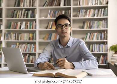 Portrait of serious diligent Indian student guy in glasses make exercise, writes summary studying using laptop seated at desk with textbooks and copybook, look at camera. Higher education, development - Powered by Shutterstock - Get 15% off with code: PIKWIZARD15