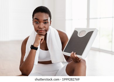 Portrait Of Serious Disappointed African American Black Woman Holding Bathroom Scales Wearing White Sportswear And Towel After Workout. Lady Not Satisfied About Her Weight Gain, Looking At Camera