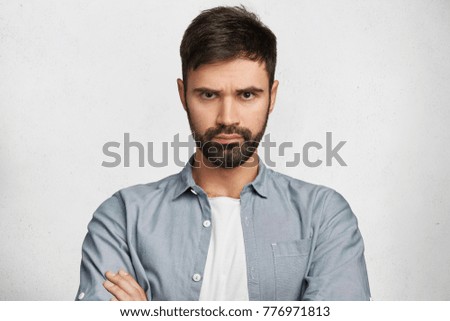Portrait of serious displeased male stands crossed hands against white background, scowl face in dissatisfaction, has quarrel with wife. Discontent man has stubble, expresses annoyannce and negativity