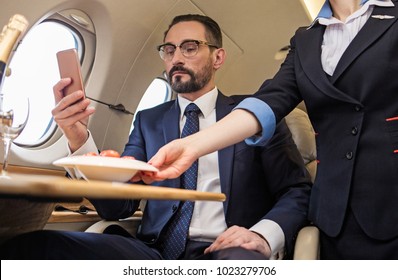 Portrait Of Serious Elegant Male Looking At Mobile Phone While Sitting At Tray Table In Airplane. Stewardess Is Serving Plate Of Food. Low Angle