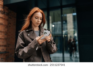 Portrait of serious fashionable young woman with red hair using smartphone standing on city street. Focused redhead female typing on smartphone outdoors on summer day. - Powered by Shutterstock