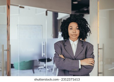 Portrait of serious female boss inside business company office, businesswoman crossed arms looking concentrated at camera, wearing shirt, satisfied and successful woman with curly hair.