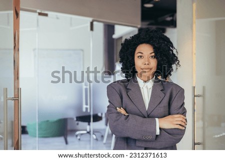 Portrait of serious female boss inside business company office, businesswoman crossed arms looking concentrated at camera, wearing shirt, satisfied and successful woman with curly hair.