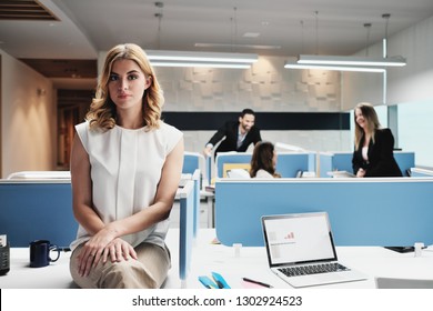 Portrait Of Serious Female Employee Excluded By Colleagues. Blonde Businesswoman Sitting On Desk At Work In Modern Coworking Space, Looking At Camera With Sad Expression.