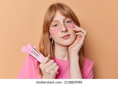 Portrait of serious ginger teenagge girl looks directly at camera applies hydrogel patches under eyes to reduce pufiness holds cosmetic brushes for doing makeup isolated over brown background