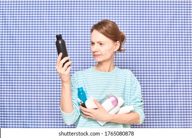 Portrait of serious girl with anti-acne skincare product on face, holding many cosmetics, reading list of ingredients on cosmetic bottle, over shower curtain background. Care for skin. Beauty concept