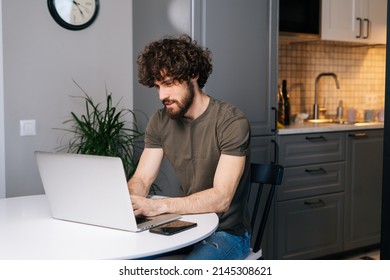 Portrait Of Serious Handsome Bearded Young Business Man Working On Project At Laptop Typing, Using Online App Sitting At Table In Kitchen Room With Modern Interior, Looking On Computer Screen.