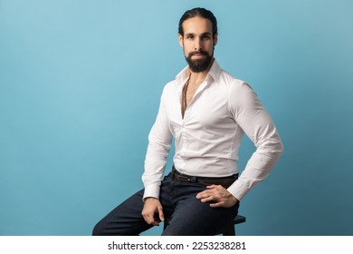 Portrait of serious handsome man with beard and collected dark wearing white shirt looking at camera with confident facial expression. Indoor studio shot isolated on blue background. - Shutterstock ID 2253238281