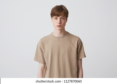 Portrait Of Serious Handsome Young Man Student With Short Haircut And Blond Hair In Beige T Shirt Standing And Looking Directly In Camera Isolated Over White Background Feels Concentrated