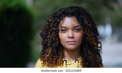 Portrait Of A Serious Hispanic Latina Girl With Curly Hair Standing Outside Looking At Camera. African American Young Millennial Woman In 30s Wearing Yellow Blouse