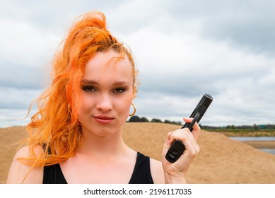 Portrait Of A Serious Insidious Secret Agent Girl In A Black Dress With Red Hair With A Gun In Her Hands Against The Background Of A Sandy Beach.