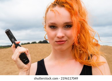 Portrait Of A Serious Insidious Secret Agent Girl In A Black Dress With Red Hair With A Gun In Her Hands Against The Background Of A Sandy Beach.