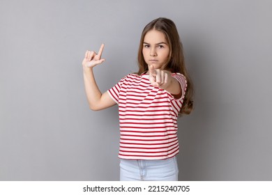 Portrait Of Serious Little Girl Wearing Striped T-shirt Showing Looser Gesture And Pointing At Camera, Looking With Grumpy Face. Indoor Studio Shot Isolated On Gray Background.