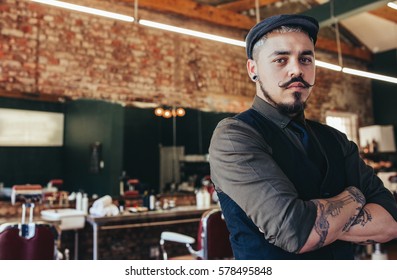 Portrait Of Serious Looking Young Man Standing With His Arms Crossed At Barber Shop. Male Hairdresser Standing In His Salon.