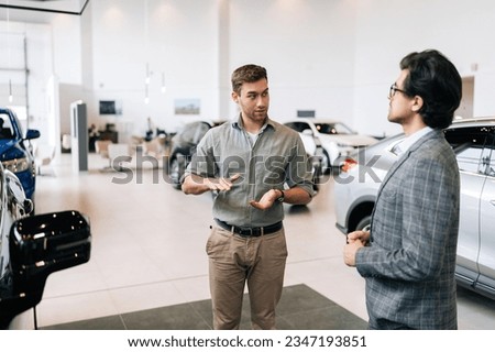 Portrait of serious male client talking to professional car dealer in business suit in dealership discussing automobiles looking at luxurious new model. Concept of choosing and buying new auto.