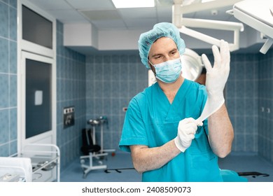 Portrait of serious male surgeon wearing medical scrubs, surgical cap and face mask putting on latex gloves in operating theatre. - Powered by Shutterstock