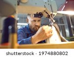 Portrait serious male worker making shoes in factory workshop. Concentrated man sewing and looking at needle sitting at table with sewing machine. Footwear and clothes manufacturing industry concept