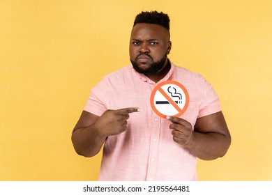 Portrait of serious man wearing pink shirt holding and pointing index finger at no smoking sign in hands, quit smoking, no tobacco day. Indoor studio shot isolated on yellow background. - Powered by Shutterstock