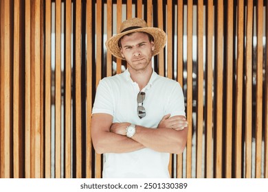 Portrait of a serious man in a white polo shirt and straw hat standing with crossed arms against a wooden slat wall background. The man looks directly at the camera with a confident expression - Powered by Shutterstock