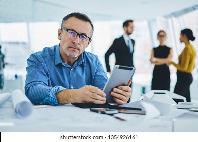 Portrait Of Serious Mature 50 Years Old Businessman In Eyeglasses For Vision Correction Looking At Camera While Sitting At Office Desktop With Digital Tablet, Concept Of Management