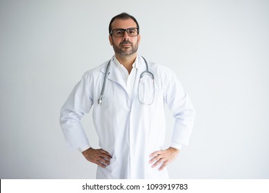 Portrait Of Serious Mid Adult General Practitioner In Glasses. Caucasian Doctor Wearing Lab Coat And Stethoscope Standing With Hands On Hips. Medical Staff Concept